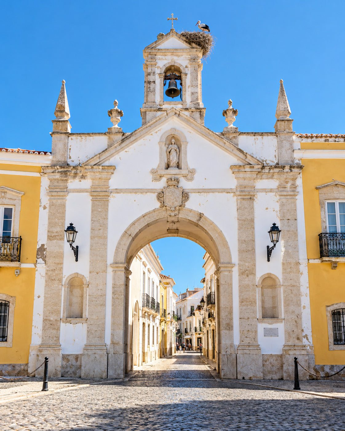 Faro: Arco da Vila city gate, pale yellow walls, cobbled street and a stork nest in sunshine